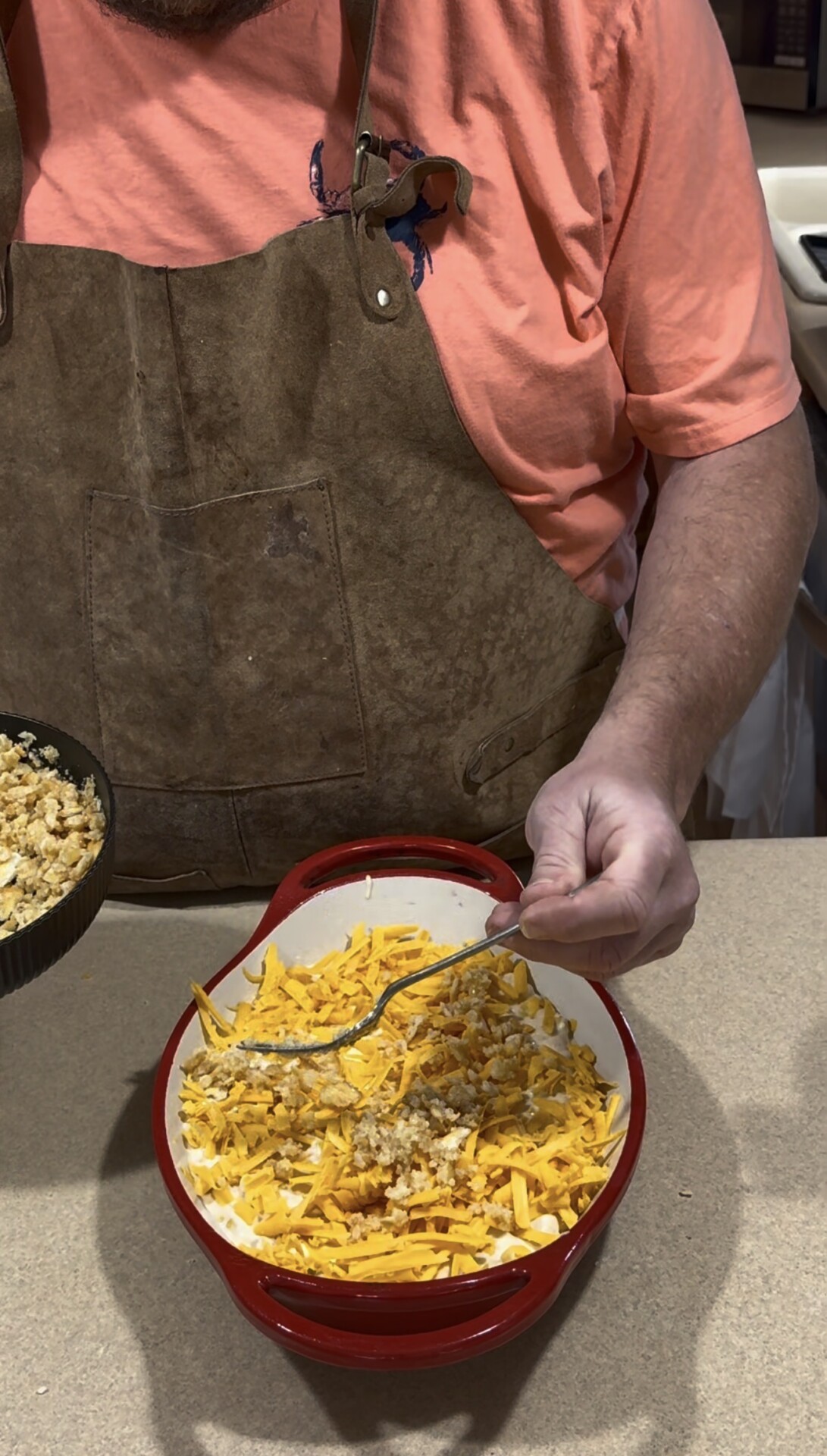 A person wearing a brown apron and orange shirt is sprinkling a breadcrumb mixture from a pan over shredded cheese in a red baking dish on a kitchen counter.