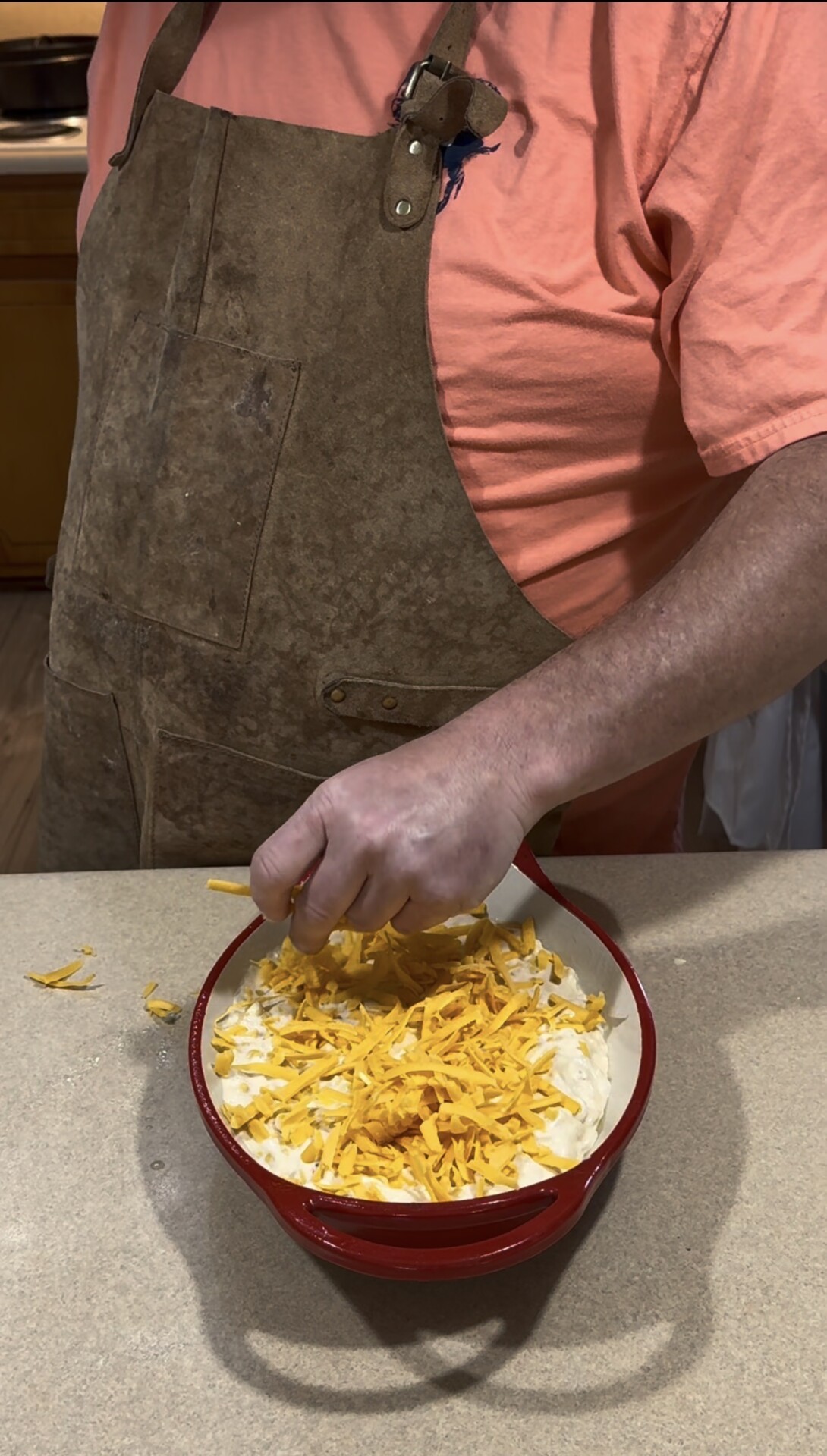 A person wearing a brown apron and orange shirt is sprinkling shredded cheddar cheese over a dish of sliced potatoes on a kitchen counter.