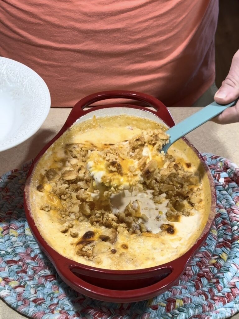 A person scoops a cheesy casserole with a crumb topping from a red baking dish into a white bowl, held over a colorful braided placemat.