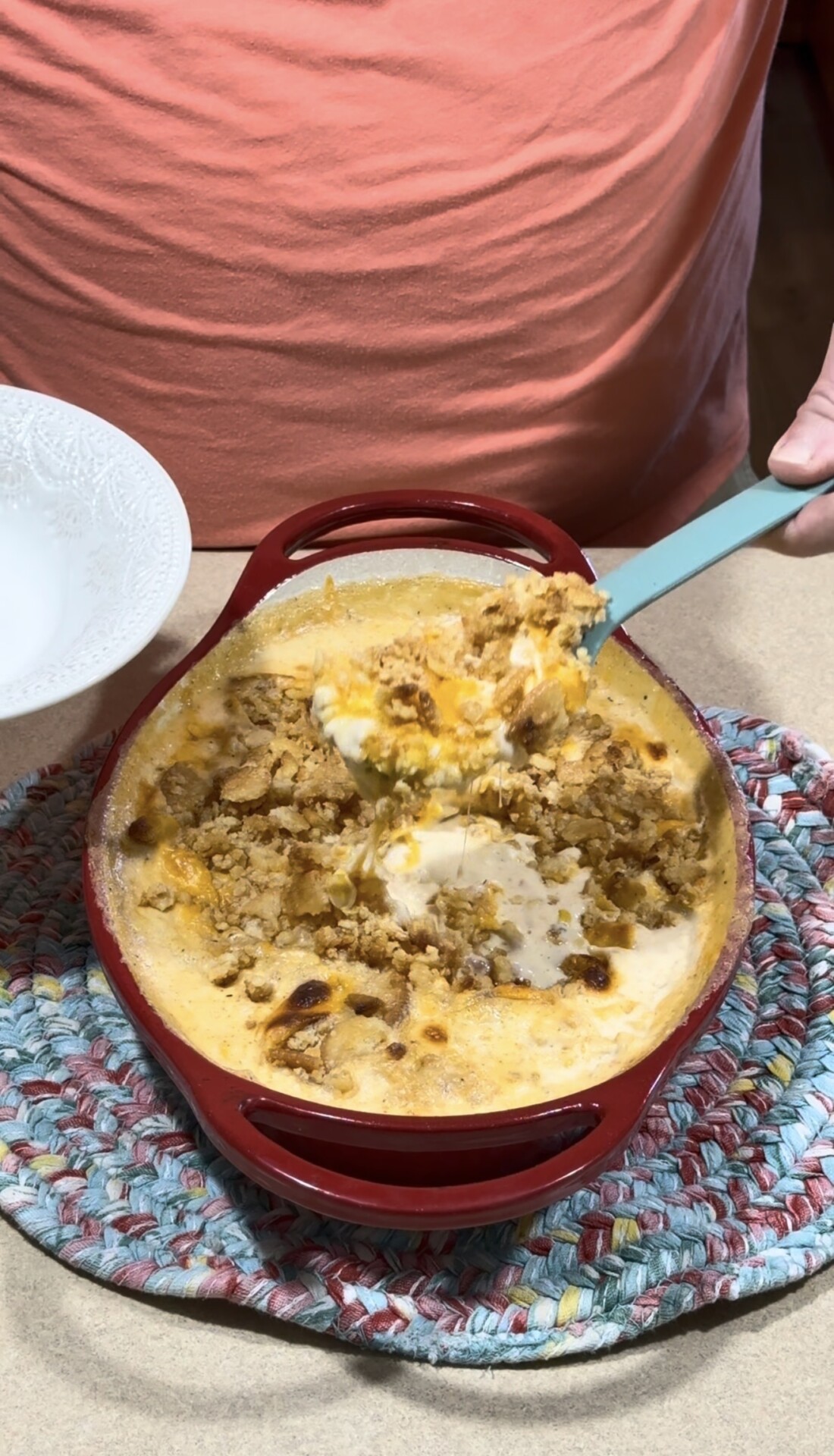 A person serves a cheesy, baked casserole from a red dish with a blue spatula onto a white plate. The casserole sits on a colorful braided trivet on a countertop.