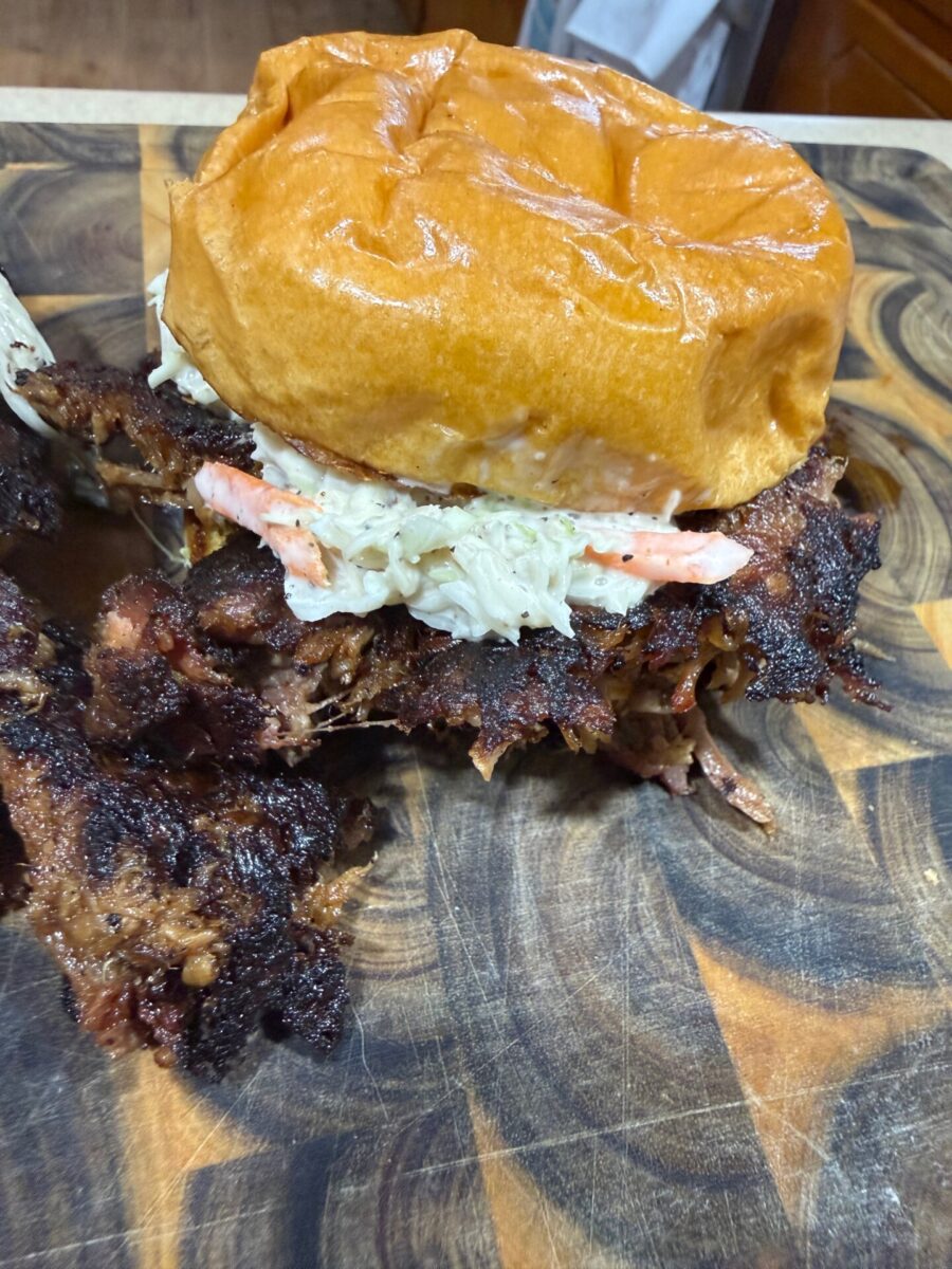 A pulled pork sandwich with coleslaw on a toasted bun sits on a wooden cutting board, with barbecue meat spilling out from the sides.