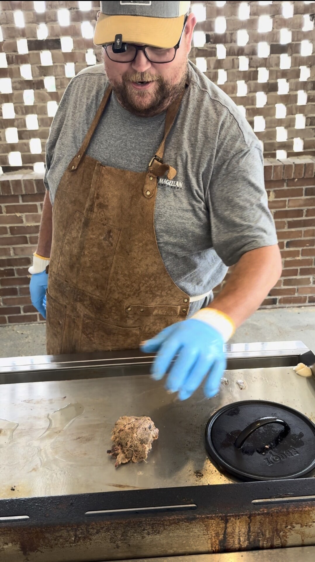 A man wearing a brown apron, gray shirt, and blue gloves cooks on a flat-top grill. He shapes a ball of ground meat next to a cast iron press, with a brick wall in the background.