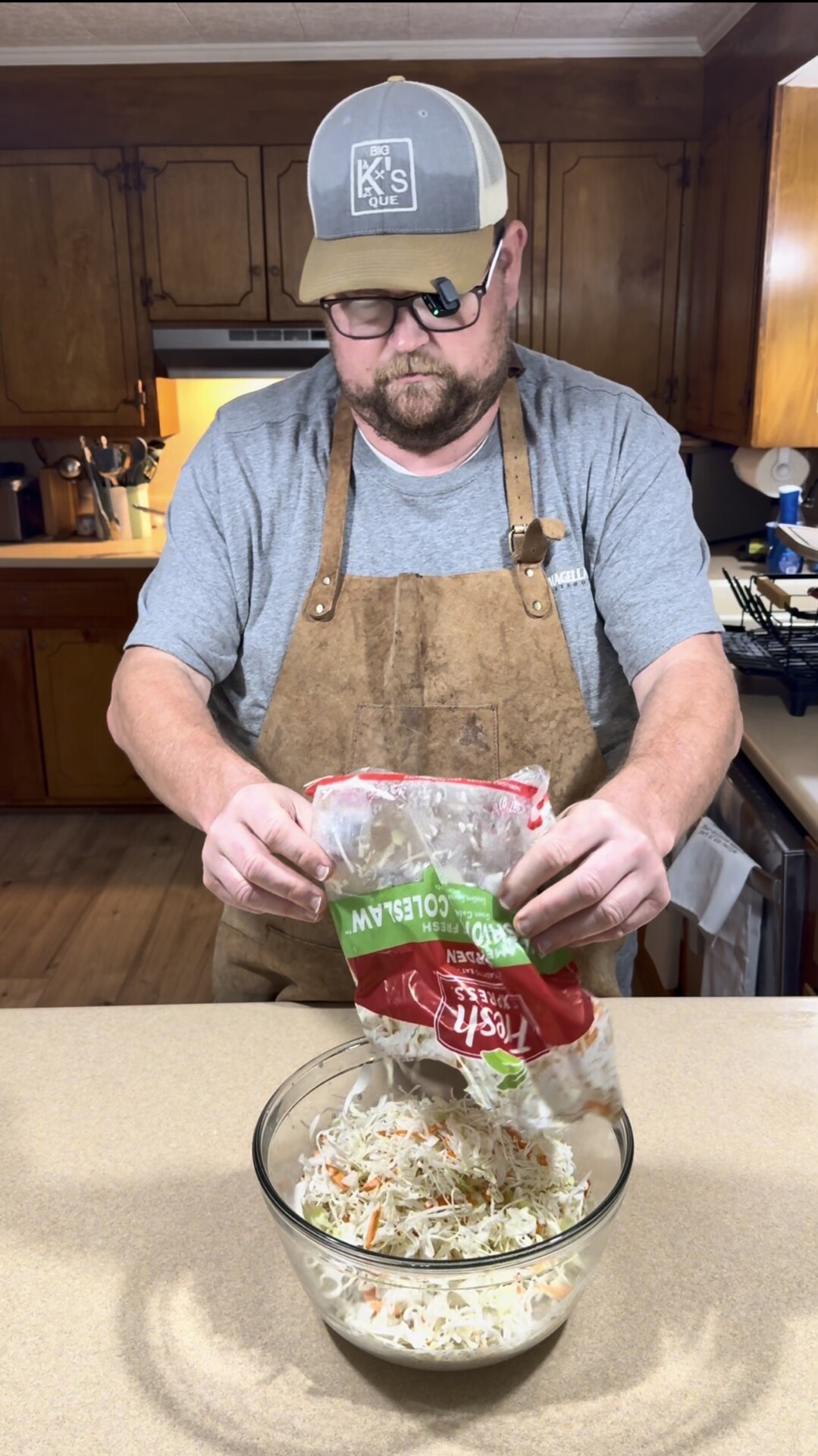 A man wearing a brown apron and cap pours bagged coleslaw mix into a glass bowl on a kitchen counter, preparing food in a home kitchen setting.