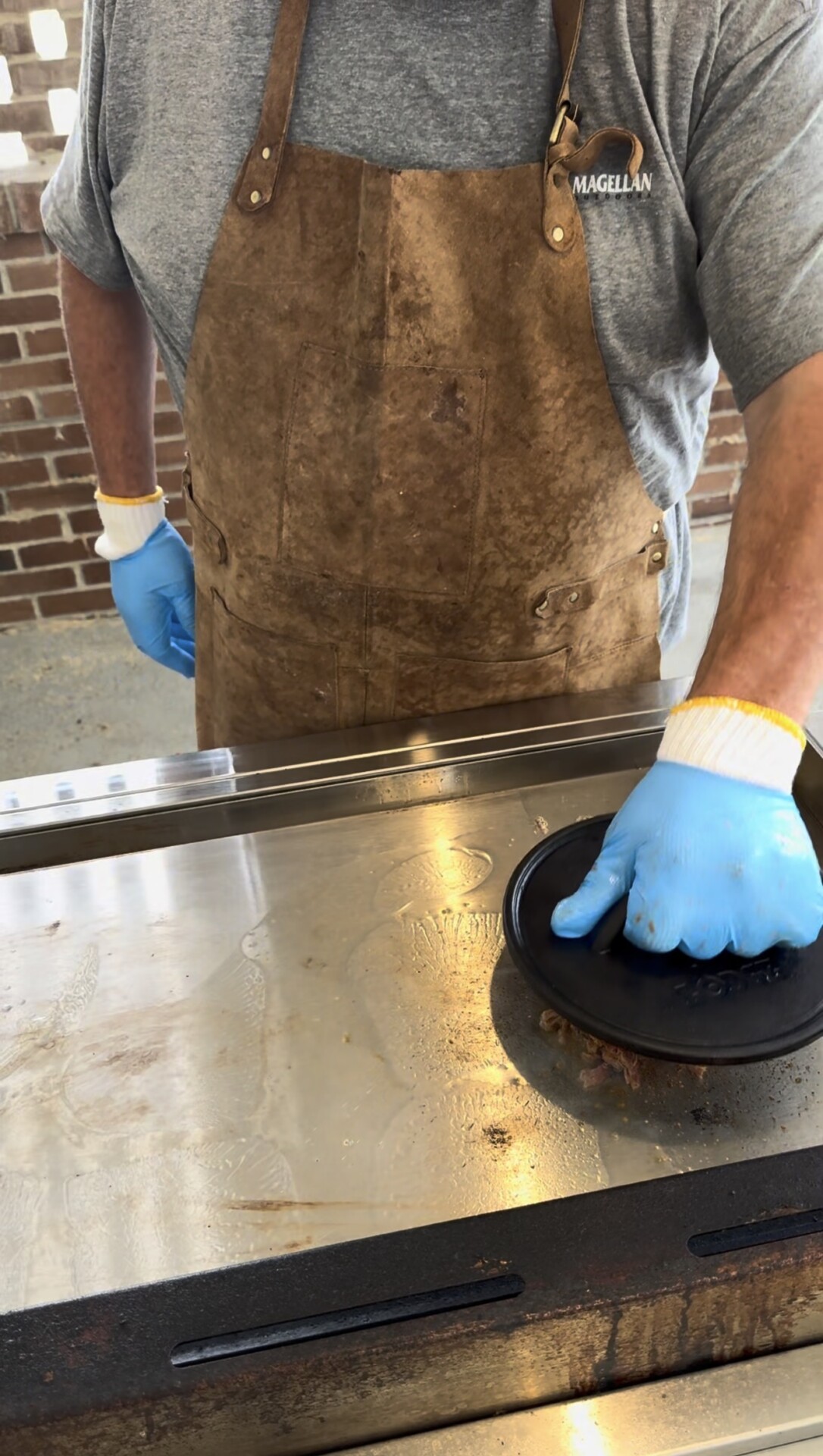 A person wearing a brown apron and blue gloves presses a metal tool onto food cooking on a flat-top grill, with a brick wall in the background.