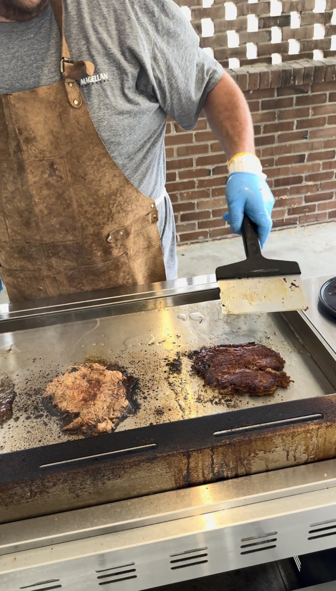 A person wearing a brown apron and blue gloves cooks two pieces of meat on a large outdoor griddle, holding a metal spatula; a brick wall is in the background.