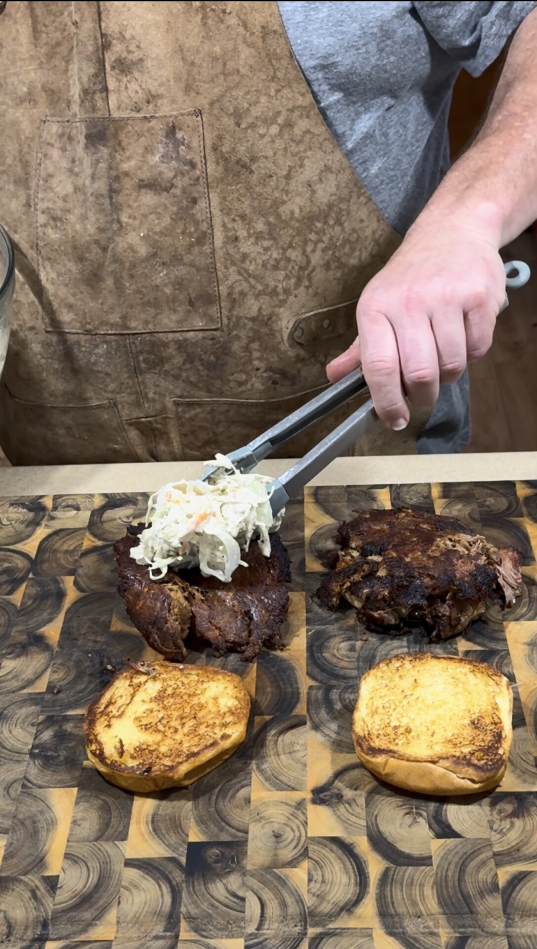 A person wearing a brown apron uses tongs to place coleslaw onto a piece of cooked meat on a toasted sandwich bun, preparing sandwiches on a patterned wooden cutting board.