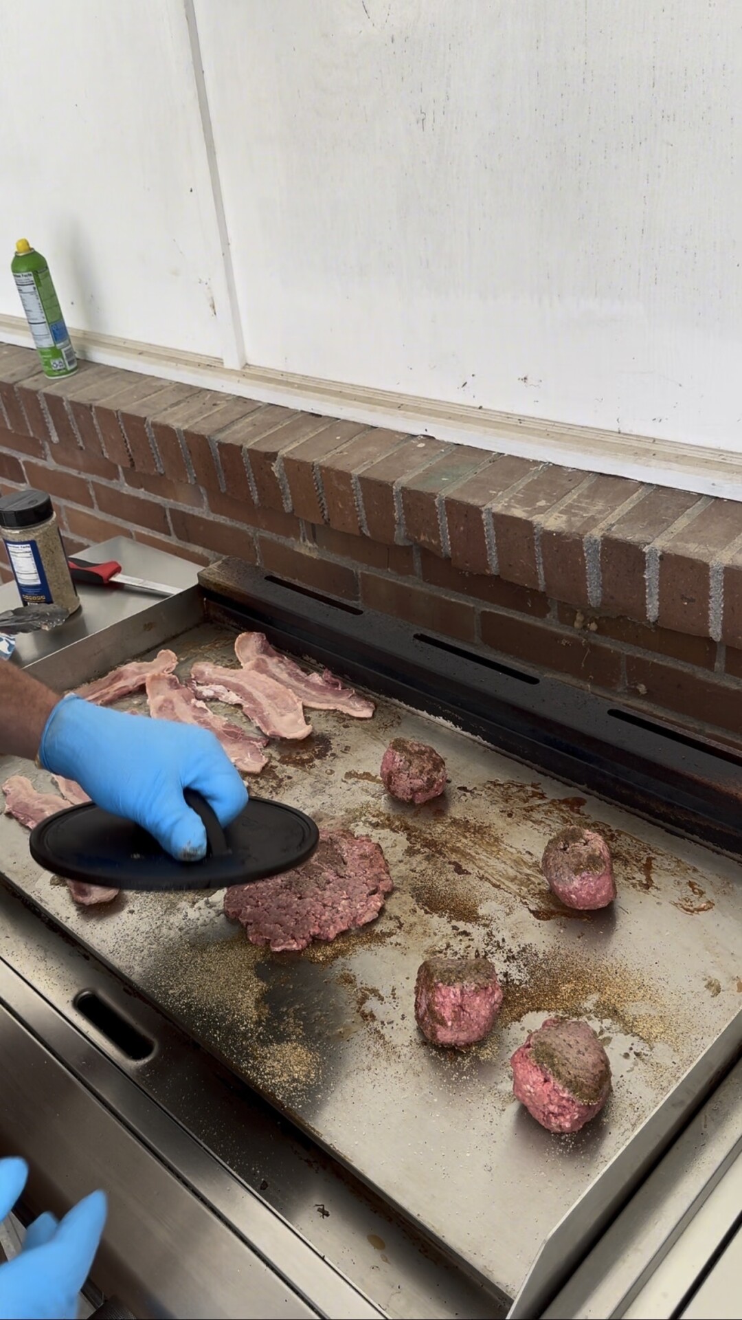 A gloved hand uses a burger press to flatten raw patties on a hot griddle with bacon strips.