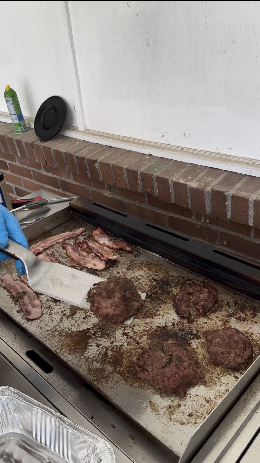 A person wearing blue gloves flips bacon and burgers on a griddle. A foil pan sits below the griddle.