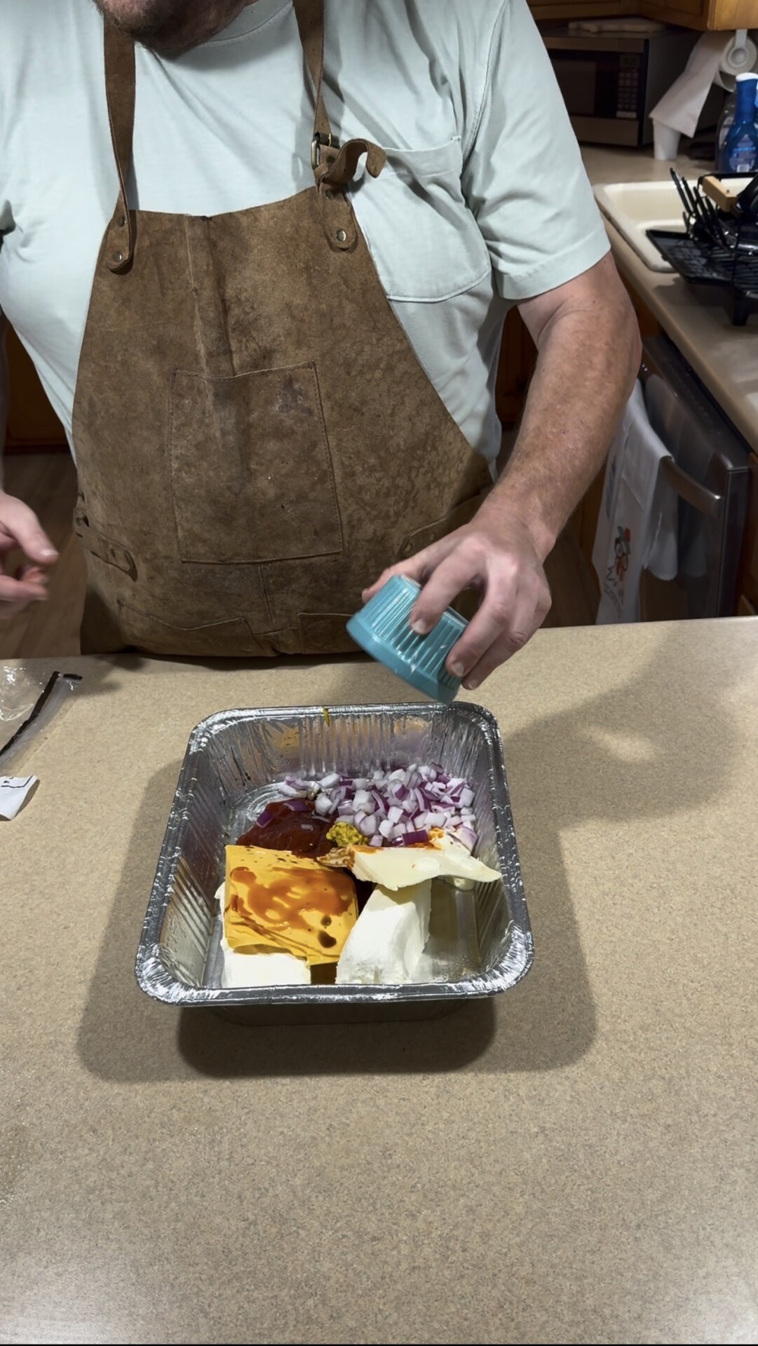 A person in a brown apron adds chopped onions to a foil pan containing cheese, BBQ sauce, and mustard.