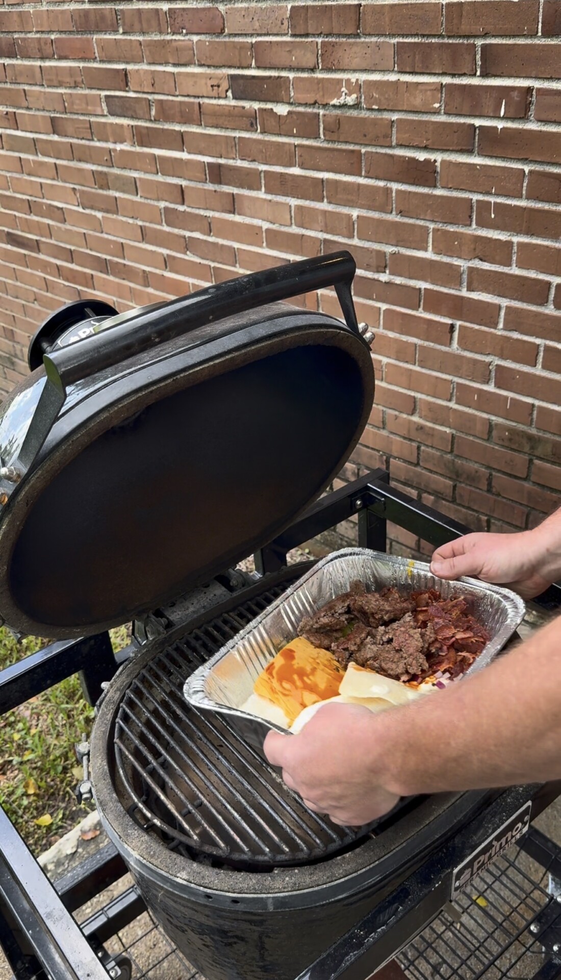 A person places a foil tray of cooked meats and cheeses on a grill with a brick wall in the background.
