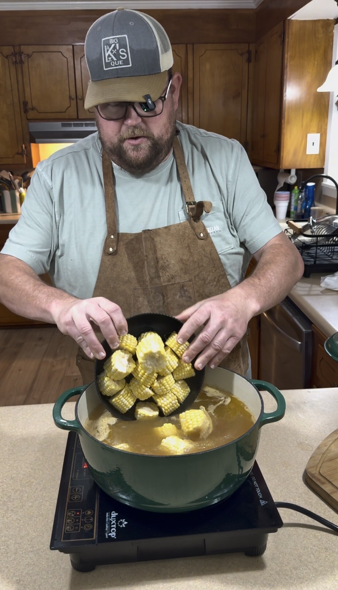 A man wearing a cap, glasses, and apron pours chopped corn on the cob into a green pot of broth on a kitchen counter, preparing a meal. Wooden cabinets and kitchen items are visible in the background.