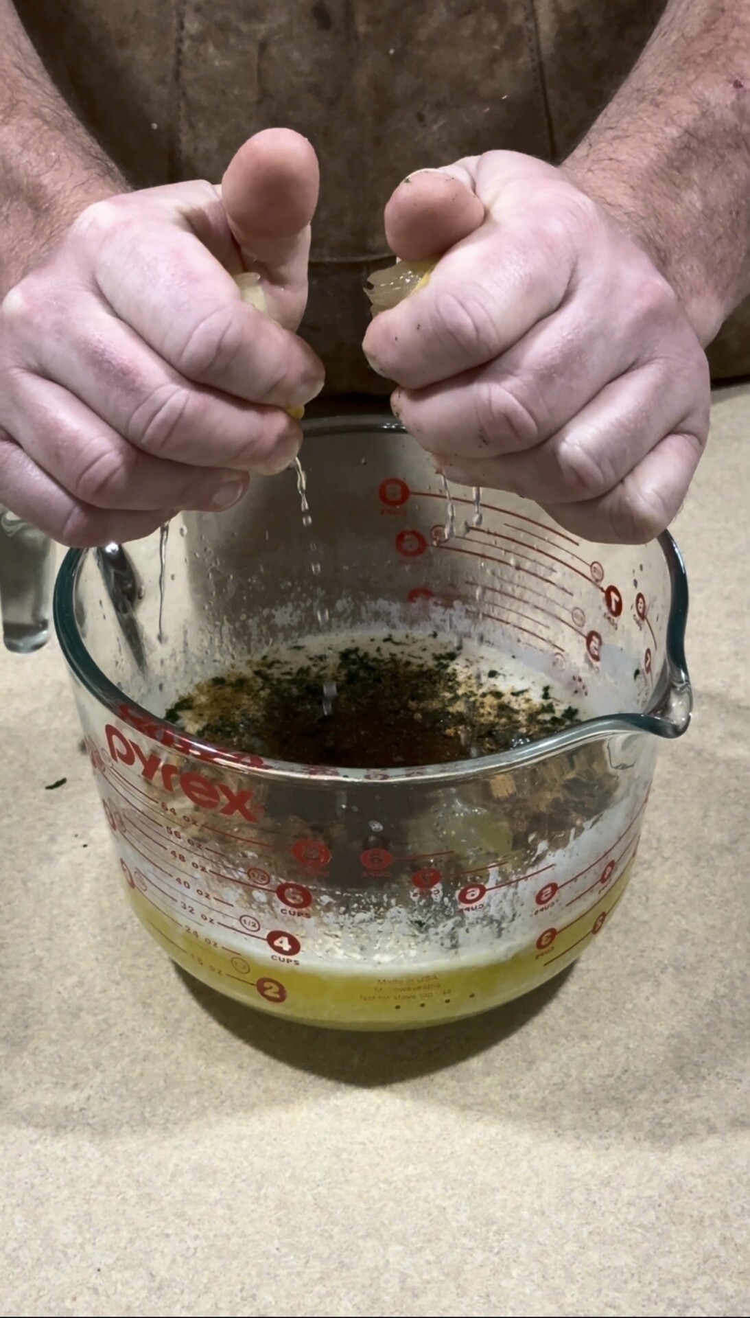 A person squeezes lemon juice into a large glass measuring cup filled with oil and spices, preparing a mixture on a kitchen counter.