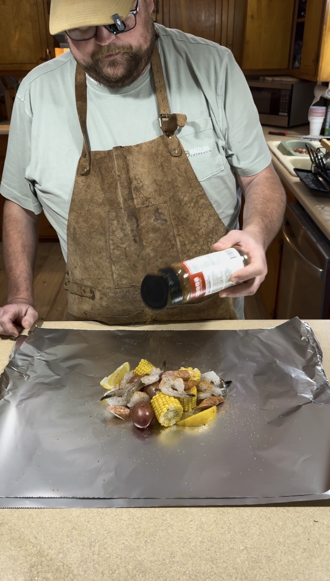 A person wearing a light green shirt and brown apron sprinkles seasoning onto a pile of shrimp, sausage, corn, and potatoes placed on a sheet of aluminum foil on a kitchen counter.