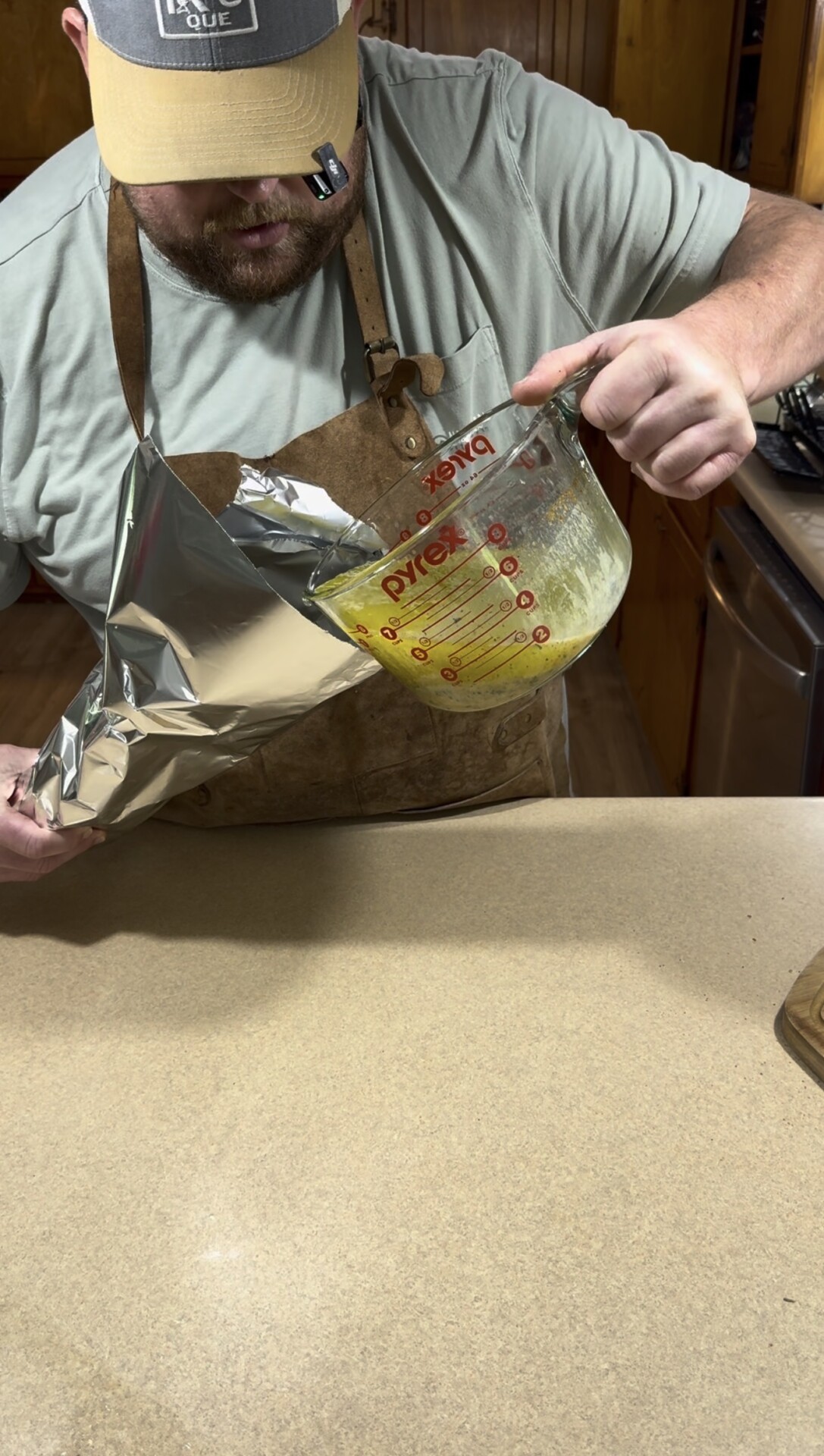 A person wearing an apron pours melted butter from a Pyrex measuring cup onto a sheet of aluminum foil over a kitchen counter.