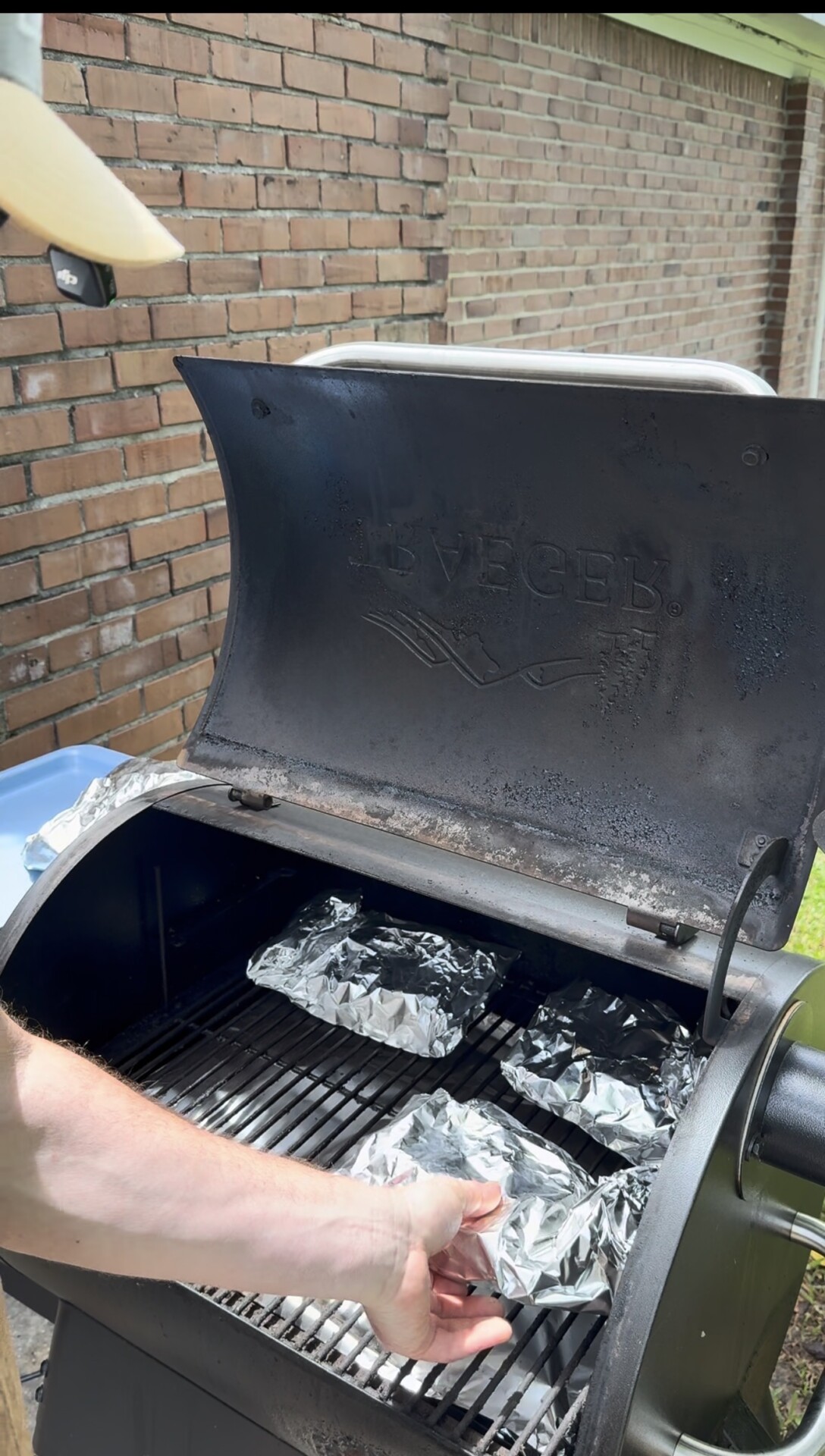 A person places a foil-wrapped item on the grill inside a Traeger smoker. Several foil-wrapped items are already on the grill, and the smoker lid is open. A brick wall and grass are visible in the background.