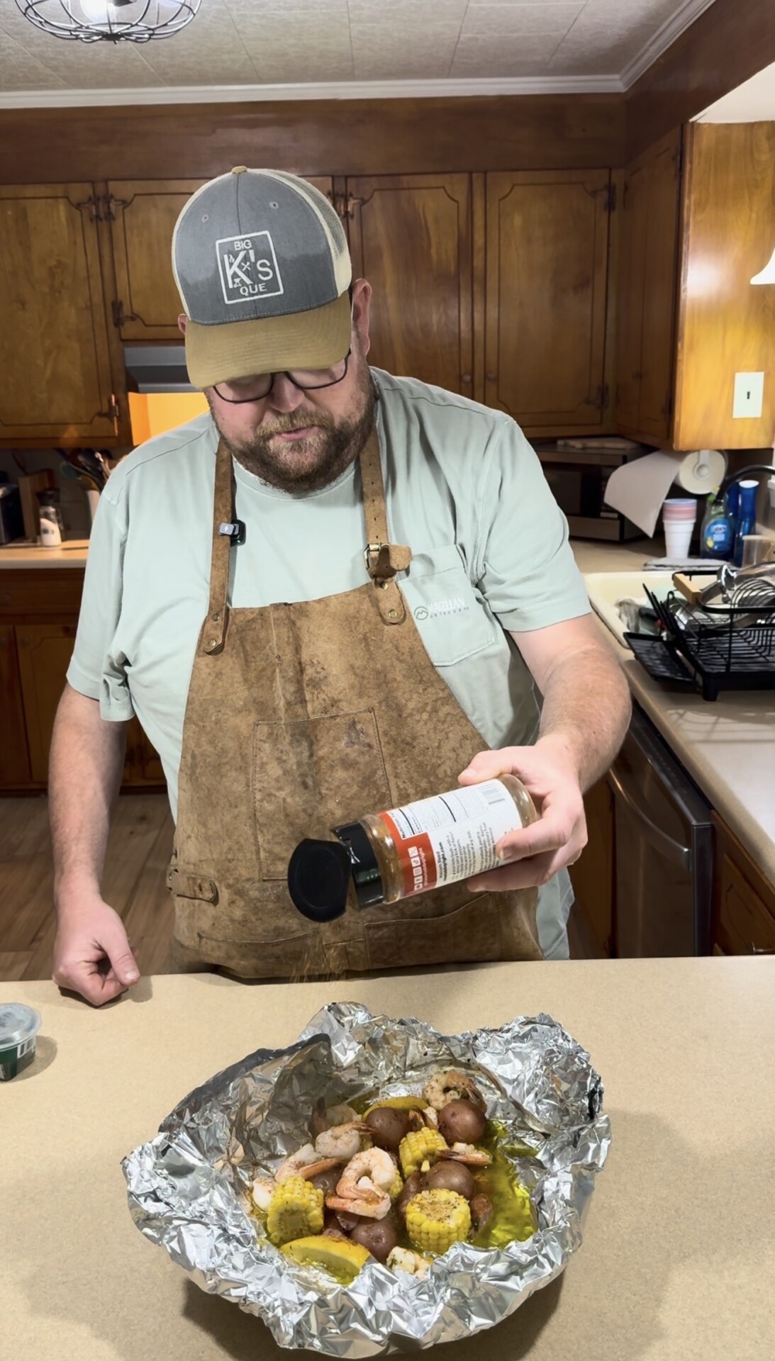 A man in an apron and cap sprinkles seasoning from a jar onto shrimp, corn, and potatoes in foil on a kitchen counter. Wooden cabinets, a dishwasher, and kitchen items are visible in the background.