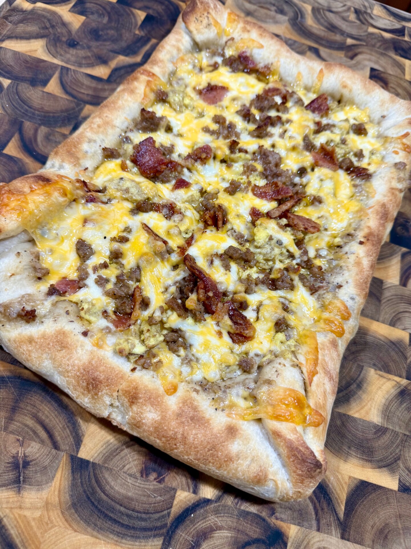 A rectangular flatbread pizza topped with melted cheese, crumbled sausage, and bacon sits on a wooden cutting board with a swirling grain pattern.