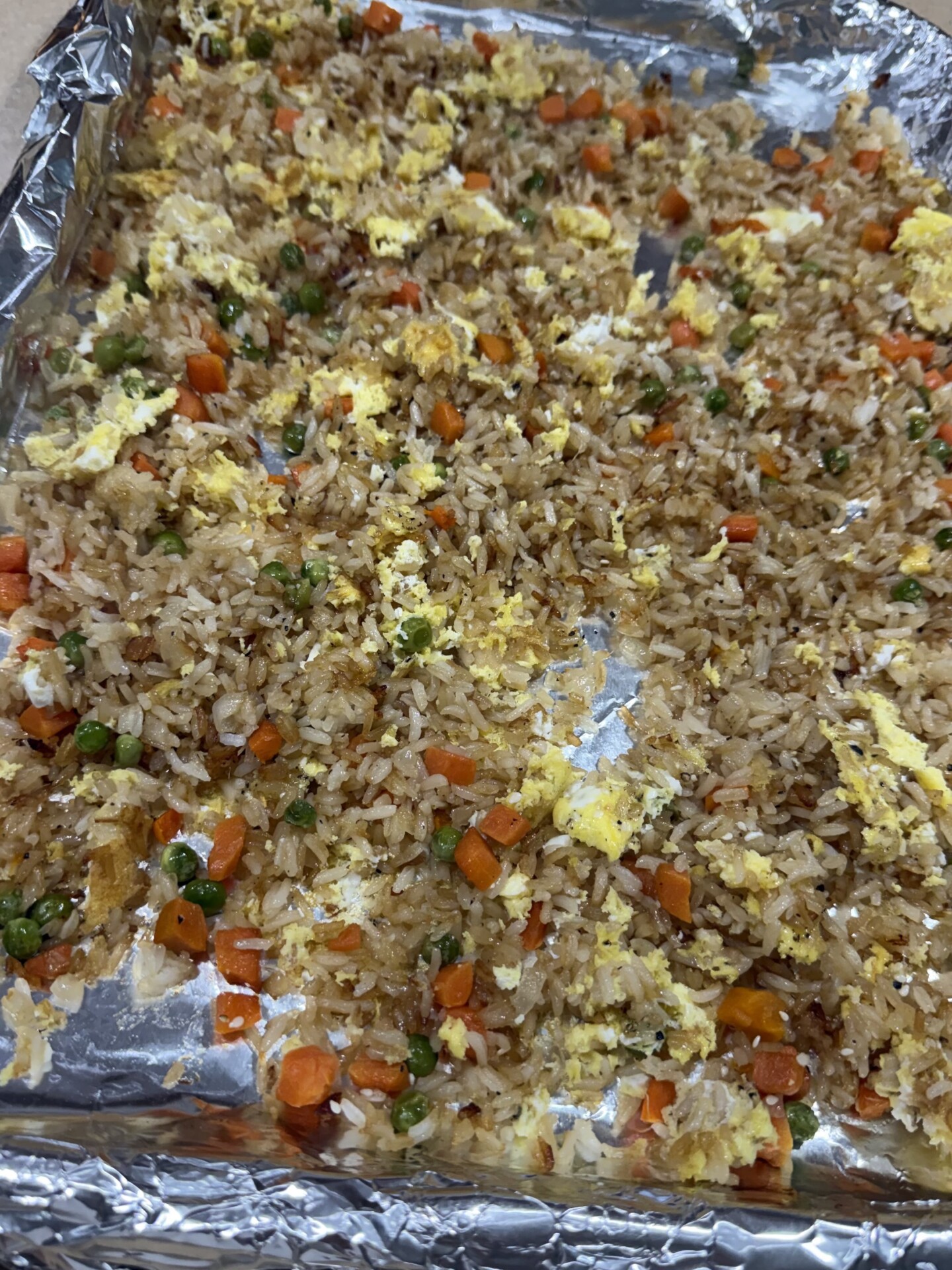 Fried rice with scrambled eggs, peas, and diced carrots scattered on a foil-lined baking sheet.