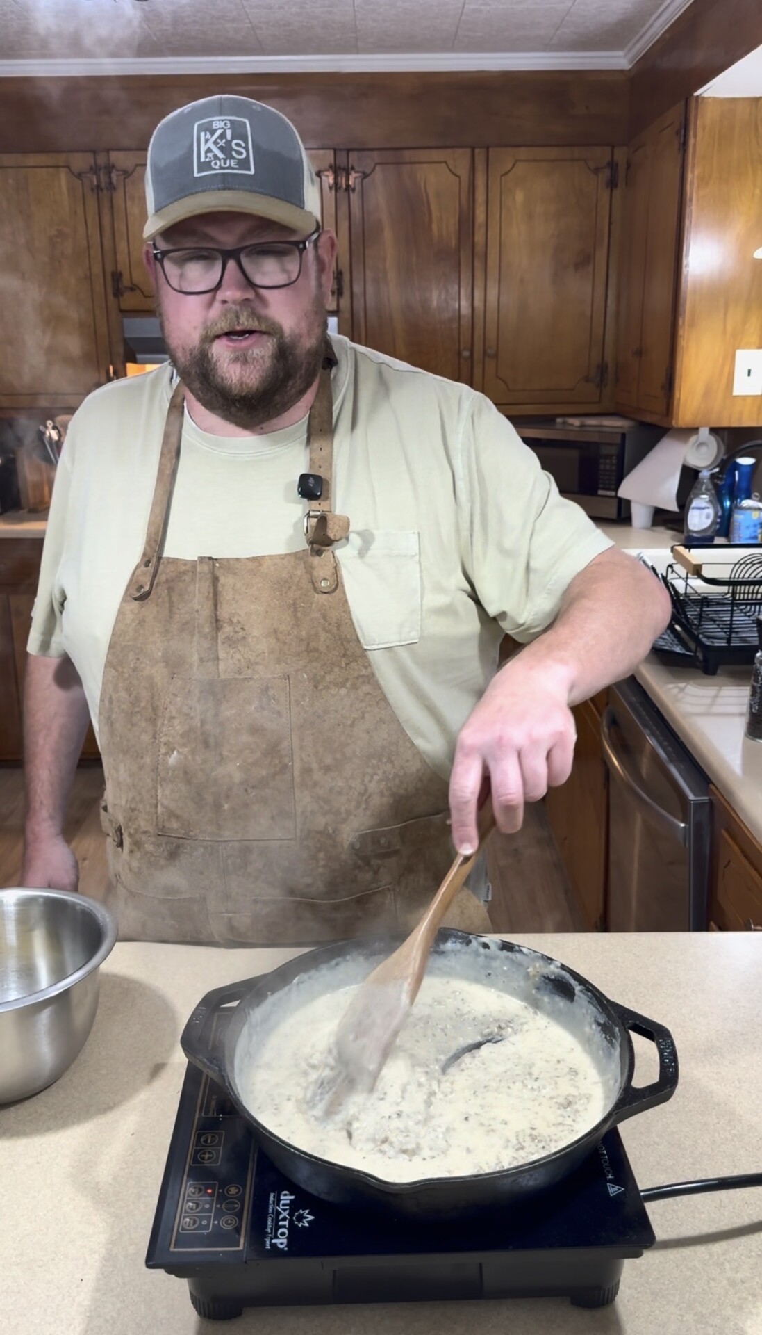 A man wearing glasses, a hat, and a tan apron stirs a creamy dish in a cast iron skillet on a stovetop in a kitchen. Wooden cabinets and various kitchen items are visible in the background.