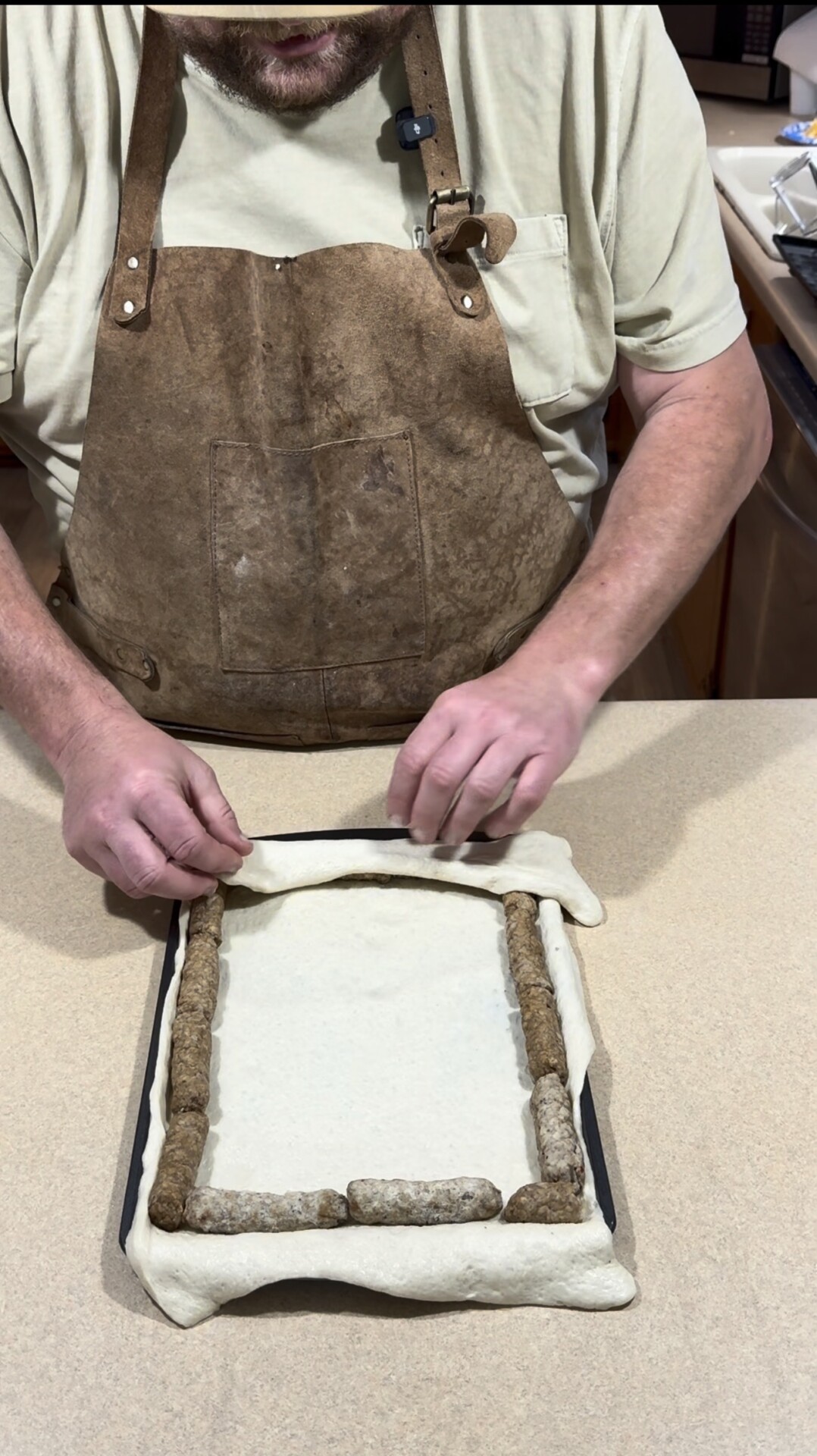 A person wearing a brown apron is folding the edge of a sheet of dough around a rectangular baking tray lined with sausage links, preparing a dish on a kitchen counter.
