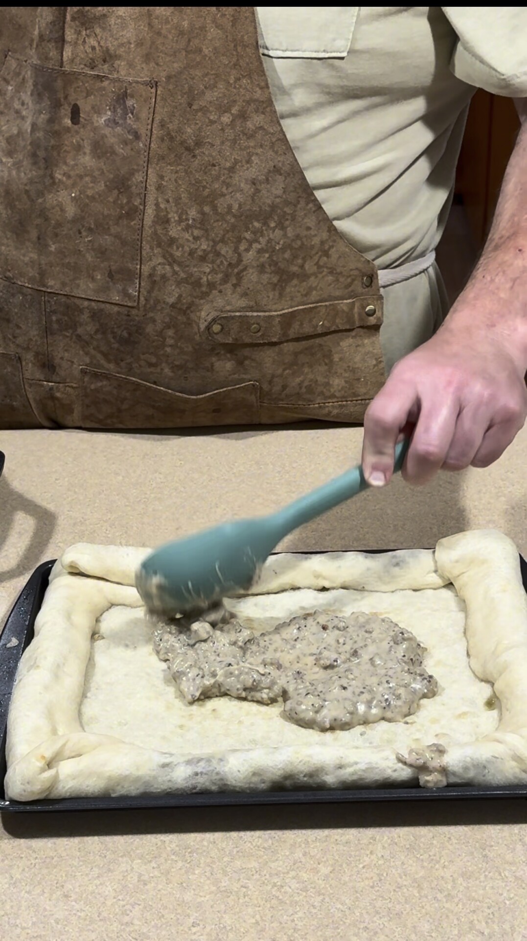 A person wearing a brown apron spreads a creamy, chunky filling onto a rectangular dough with raised edges using a blue spatula on a baking sheet.