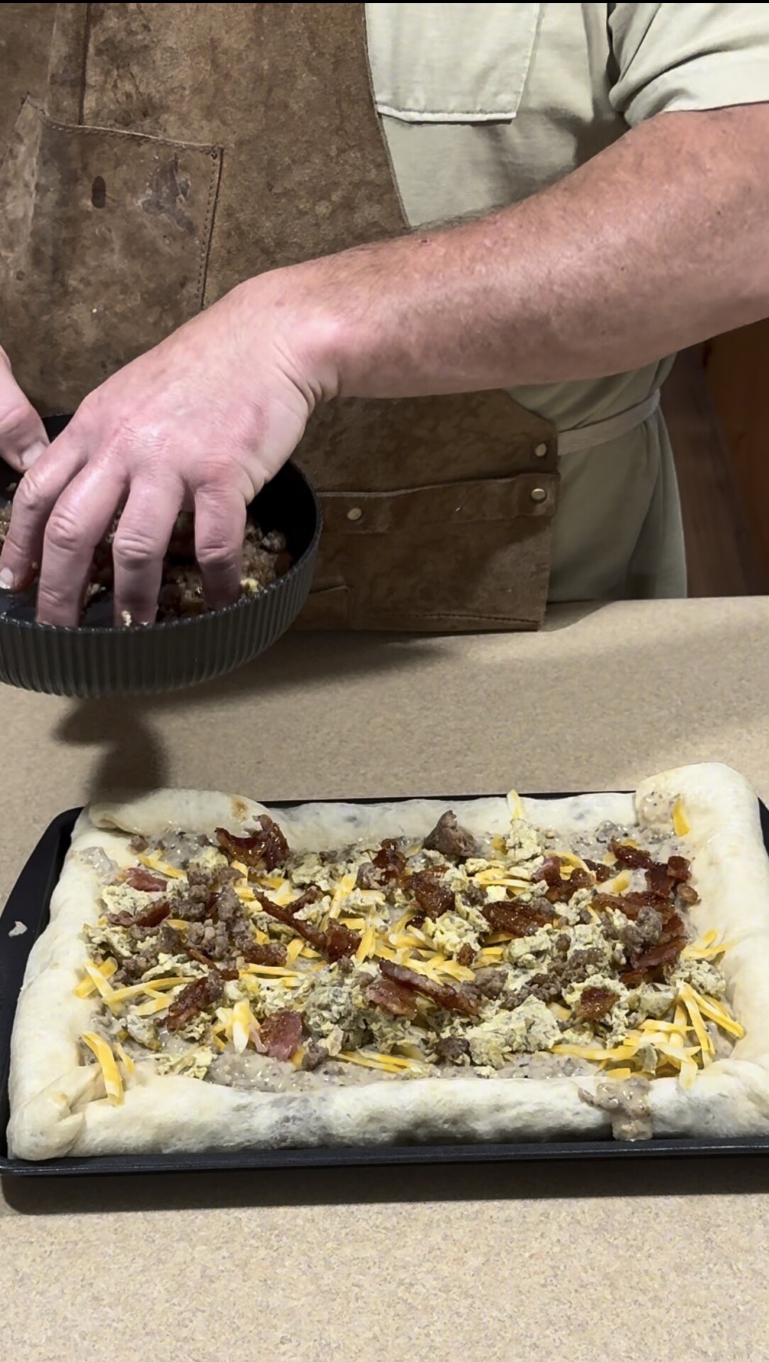 A person wearing an apron sprinkles sausage from a small pan onto an unbaked breakfast pizza topped with cheese, bacon, and sausage on a baking sheet.