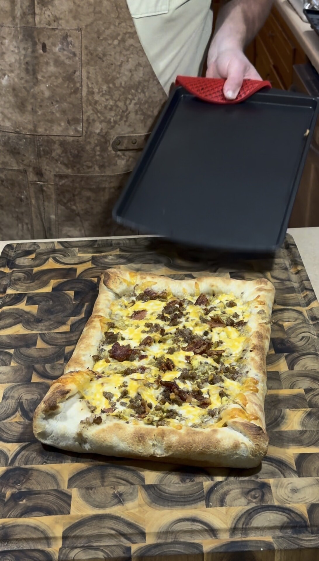 A rectangular homemade pizza topped with melted cheese and crumbled sausage sits on a patterned wooden cutting board, while a person lifts a black baking tray in the background.