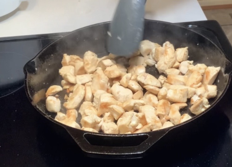 Chunks of chicken are being cooked in a cast iron skillet on a stovetop. A spatula is stirring the chicken pieces as they brown. Steam is visible, indicating the food is hot.