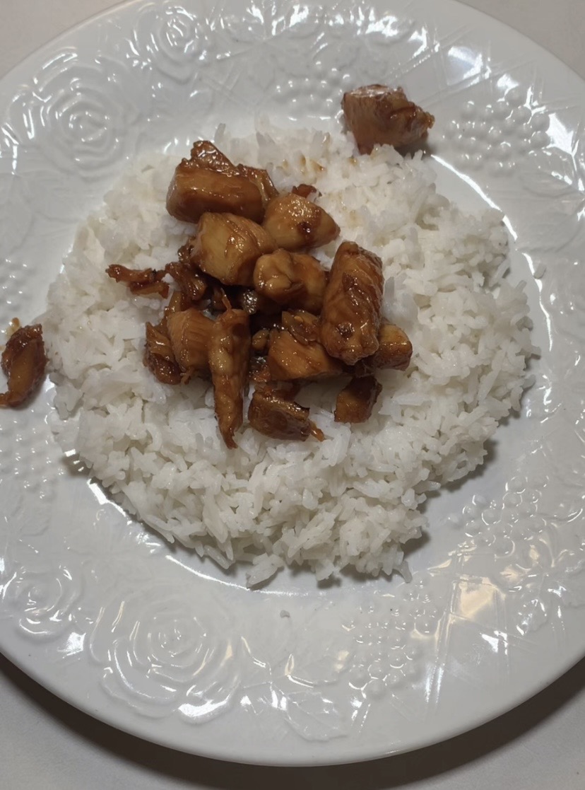 A serving of white rice topped with pieces of glazed, cooked chicken on a white floral-patterned plate.