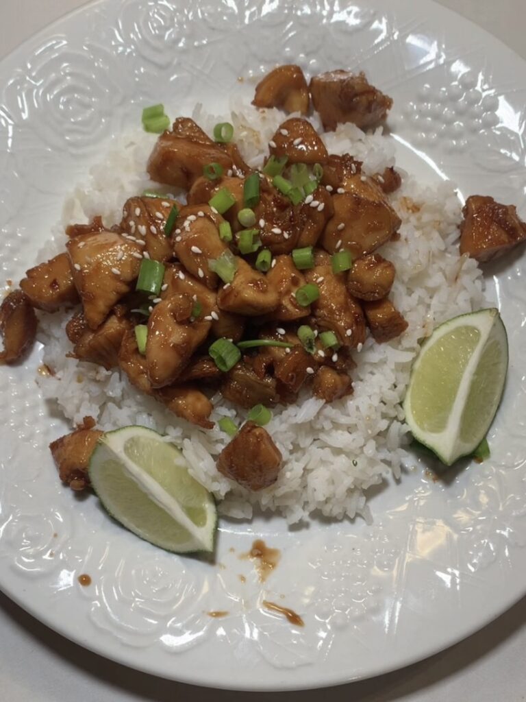 A plate of white rice topped with glazed chicken pieces, garnished with chopped green onions and sesame seeds, served with two lime wedges on the side.