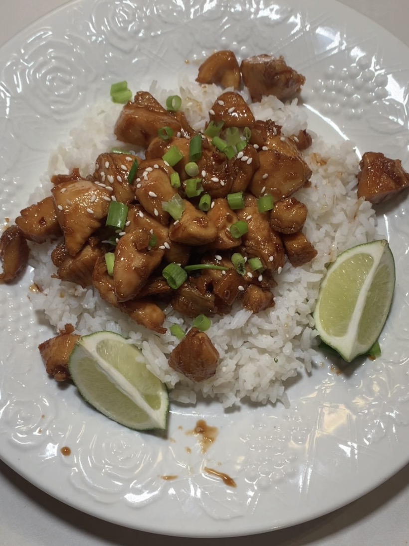 A plate of white rice topped with glazed chicken pieces, garnished with chopped green onions and sesame seeds, served with two lime wedges on the side.