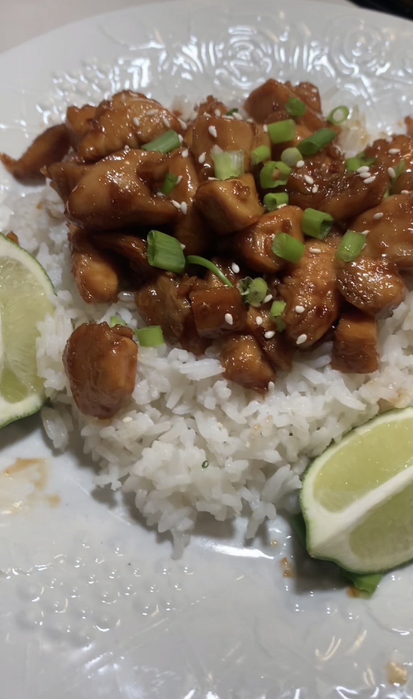 A plate of white rice topped with glazed chicken pieces, garnished with chopped green onions and sesame seeds, served with lime wedges on the side.