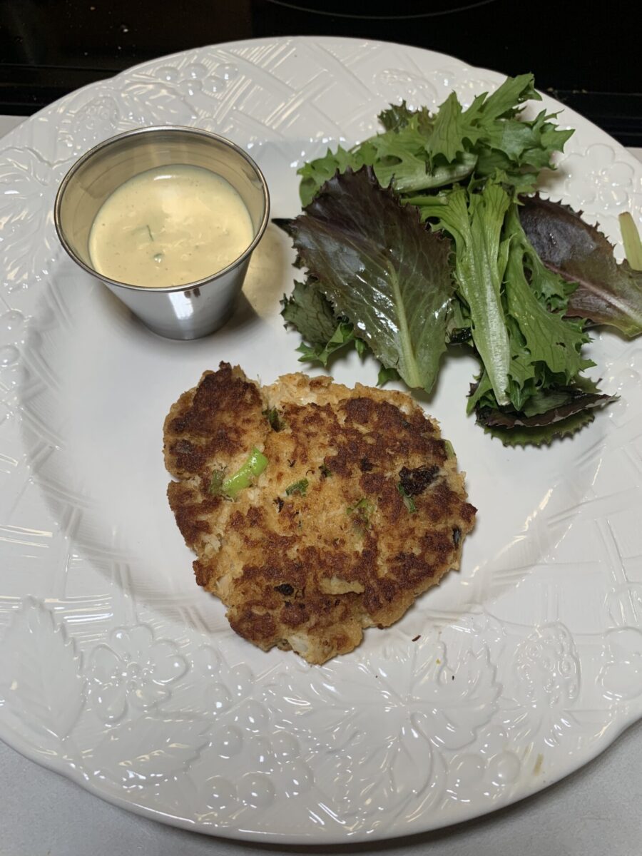 A crab cake with green herbs is served on a white plate with a small metal cup of creamy dipping sauce and a side of mixed leafy greens.