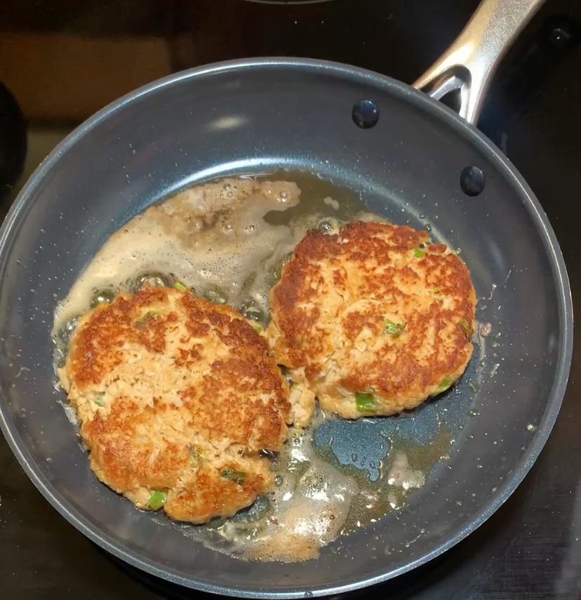 Two golden-brown patties are being cooked in a frying pan with melted butter or oil, showing crispy edges and flecks of green herbs or vegetables.