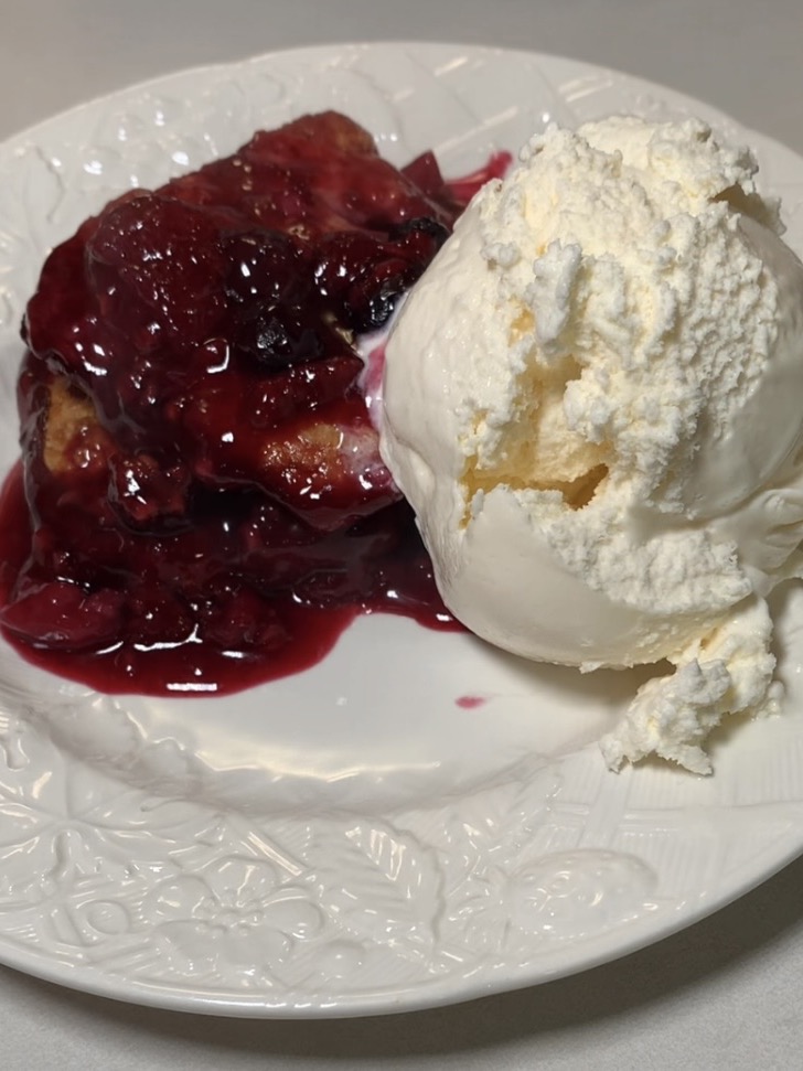 A serving of berry cobbler topped with a glossy red fruit sauce sits on a white plate next to a scoop of vanilla ice cream.