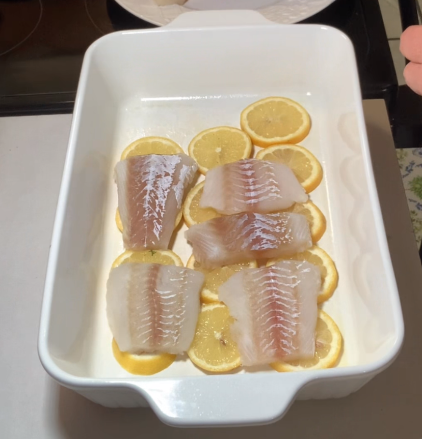 Four raw white fish fillets are arranged on top of lemon slices in a white rectangular baking dish, ready to be cooked.