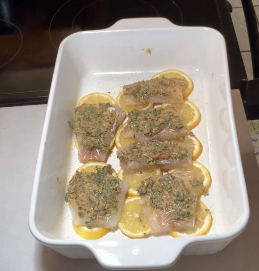 Six pieces of fish fillet topped with a green herb mixture are placed on lemon slices in a white baking dish, ready to be cooked. The dish sits on a kitchen counter next to a stovetop.