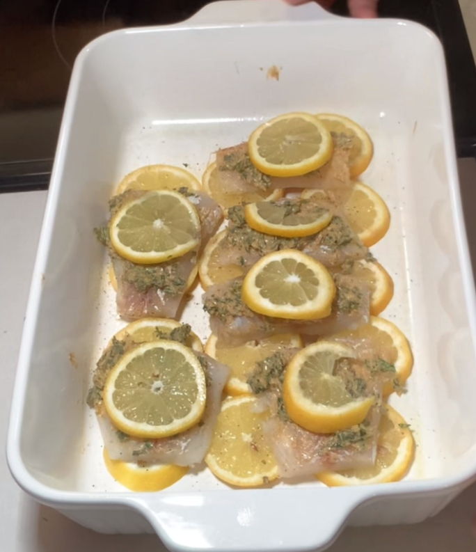 Raw fish fillets topped with herb mixture and lemon slices are arranged on a bed of lemon slices in a white baking dish, ready to be cooked.