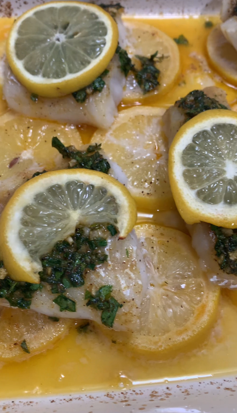 Baked fish fillets topped with chopped herbs and slices of lemon, resting on a bed of thinly sliced lemon rounds in a baking dish with visible juices.