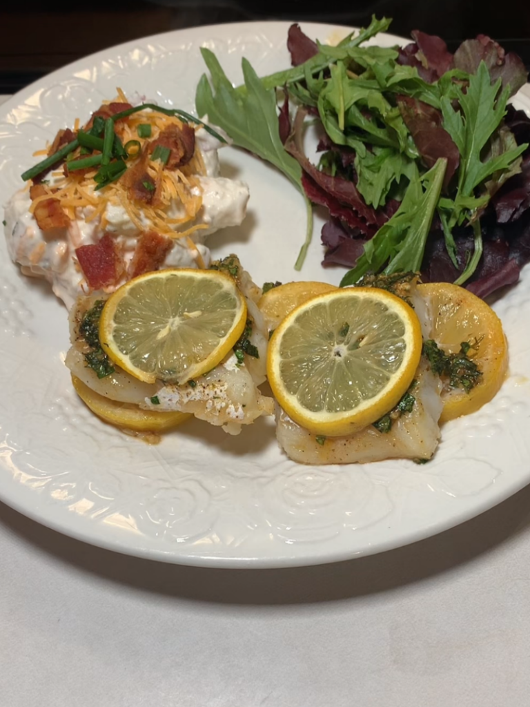 A white plate with two pieces of baked fish topped with lemon slices and herbs, a serving of loaded potato salad with bacon and chives, and mixed leafy greens.
