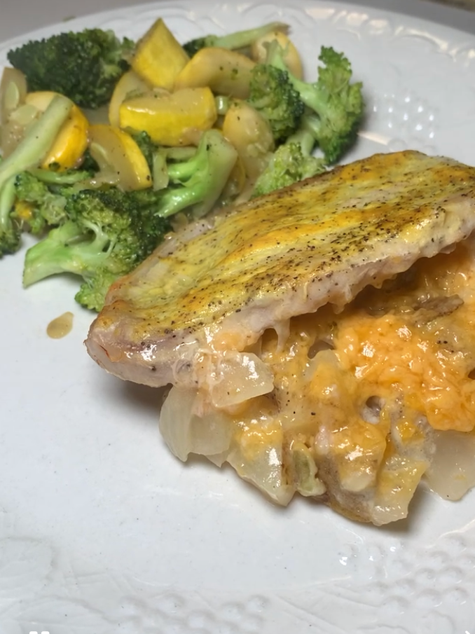 A stuffed chicken breast with melted cheese and onions is served on a white plate next to sautéed broccoli and yellow squash.