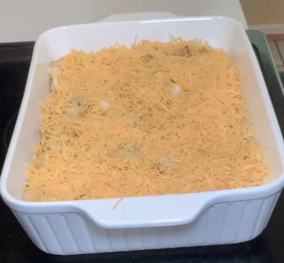 A white square baking dish filled with a casserole topped with a thick layer of shredded cheddar cheese, ready to be baked.