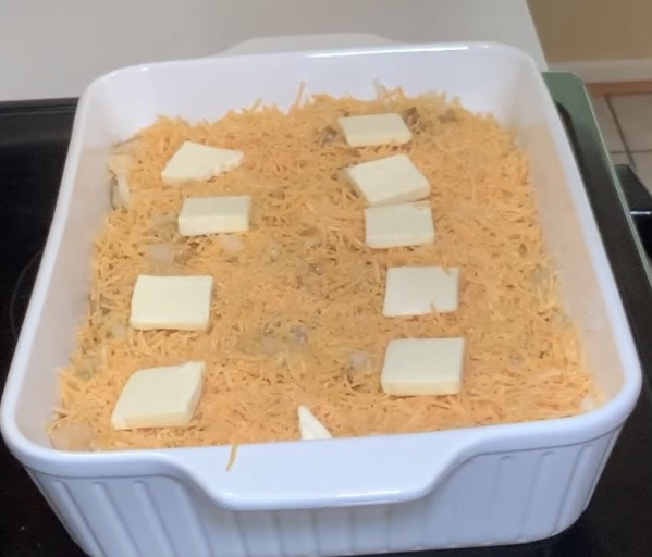 A white square baking dish filled with a casserole topped with shredded cheese and several small slices of butter, ready to be baked.