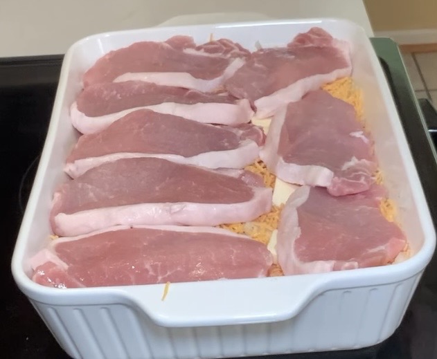 A white baking dish filled with raw pork chops arranged in a single layer on top of shredded cheese and other ingredients.