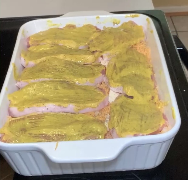 A white baking dish filled with rows of uncooked sliced meat, each piece topped with a layer of yellow mustard, sitting on a stovetop.