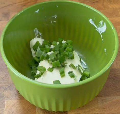 A green bowl containing a dollop of cream cheese and a handful of chopped green onions, placed on a wooden surface.