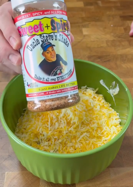 A hand holds a jar of “Uncle Steve’s Shake Sweet + Spicy” seasoning above a green bowl filled with shredded cheese on a wooden surface.