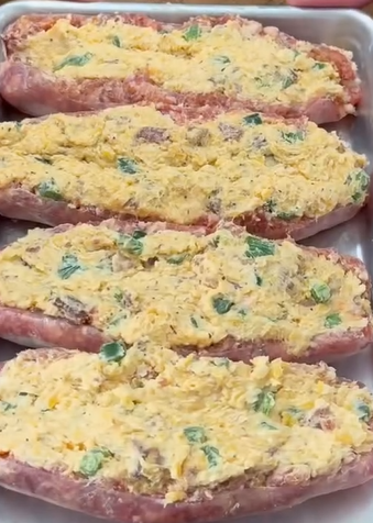 Four raw pork chops are split open and stuffed with a yellow cheesy filling mixed with green onions and possibly other ingredients, arranged in a white baking dish, ready to be cooked.