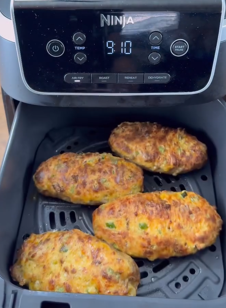 Four golden-brown stuffed potatoes sit in the basket of a Ninja air fryer, ready to serve. The air fryer display reads 9:10, and the potatoes are cooked with visible melted cheese and herbs.