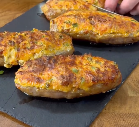Four stuffed and baked mashed potatoes with a golden, crispy cheese topping are arranged on a black slate serving tray. A hand is slicing into one of the potatoes.